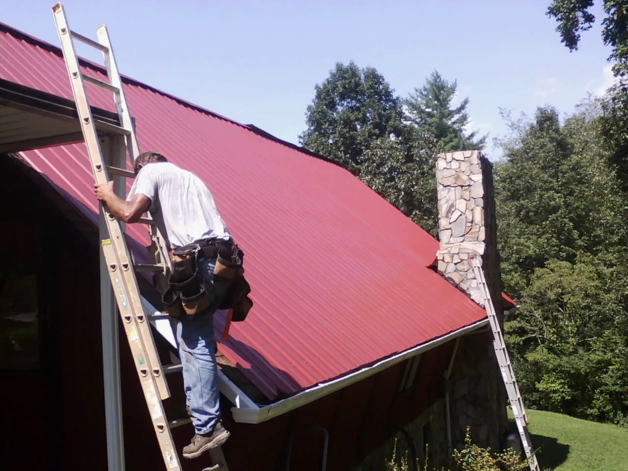 Roofer on a ladder beside a red metal roof and stone chimney — Soffit & Fascia Repair by Roofing Brothers in Shoreham